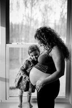 Black and white image of pregnant mom and young daughter hugging and embracing by window light