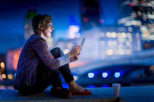 Mature businesswoman sitting on waterfront using digital tablet at night, London, UK