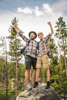 Father and son in forest standing on rock arms raised in triumph smiling, Red Lodge, Montana, USA