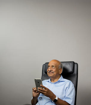 App inventor Ramesh Jain sits at his desk holding his smartphone and pointing his other hand up in the air