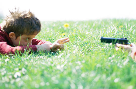 Child lying on grass pointing fingers like gun across from second person pointing gun