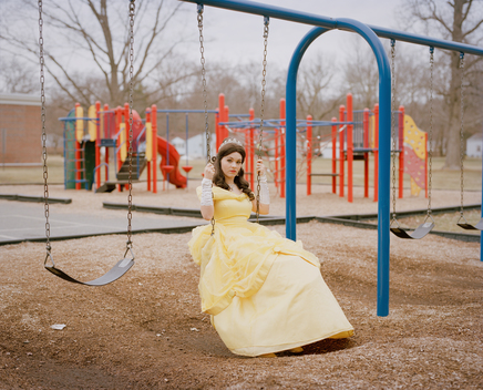 Portrait of Sapphire Nova, an ordinary woman who works as and impersonates Disney Princess Belle in yellow dress swinging on a swing set in the playground of a public school. New Jersey