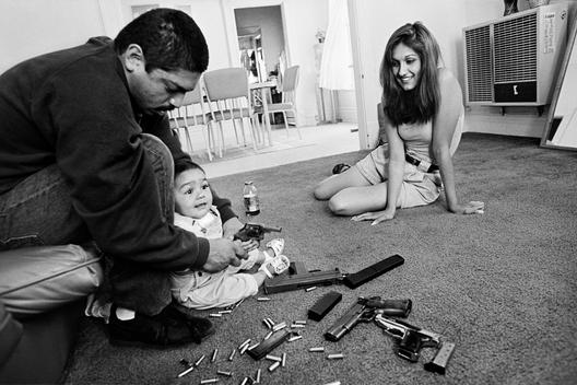 The morning after a rival gang tried to shoot Chivo for the fourth time. Chivo teaches his daughter how to hold a .32-caliber pistol. Her mother looks on. Boyle Heights. 1993