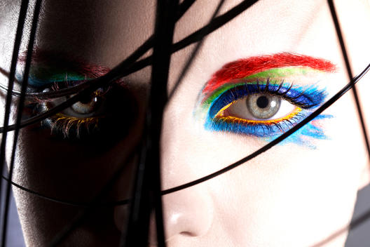 close up of the eye surrounded by black long of grass. Eye decorated with colorful eye make up, including bright red eye brows