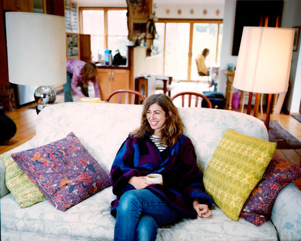 A beautiful young woman with a colorful wool coat sits on her couch amidst many colorful patterned pillows.