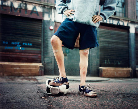 Shot Of A Young Boy, Shot Below The Waist, Hands On Hips, Standing With One Leg On A Burst Soccer Ball In An Urban Street