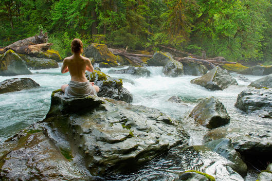 Partially clothed woman meditating on rock by water
