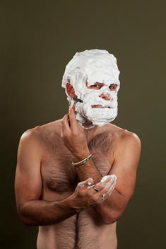 A shirtless, hairy man grimaces as he holds up a disposable razor to his face which is completely covered with shaving cream