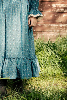 A young girl dressed in an olden day style blue dress with lace trim on the sleeve and wearing brown boots, stands in long green grass in front of a rustic wooden red brown barn wall.