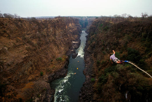 A Bungee Jumper Leaps Off The Zambezi Bridge Which Runs Between Zimbabwe (To Right Of Picture) And Zambia (To Left Of Picture) In Victoria Falls, Over The Zambezi River (Which Can Be Seen In The Distance, With White Water Rafting Taking Place On The River