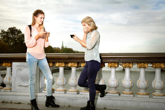 Two friends using mobile phones on Point Alexandre III bridge, laughing