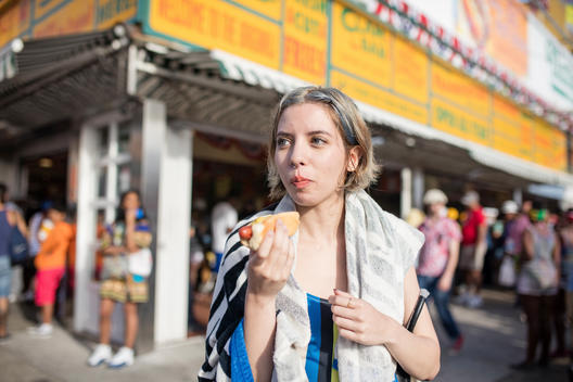 Portrait of a young Hispanic woman with a nose ring and dyed blue hair and towel over her shoulder eating a hotdog in a swimsuit outside Nathan\'s Famous in Coney Island Brooklyn, NY