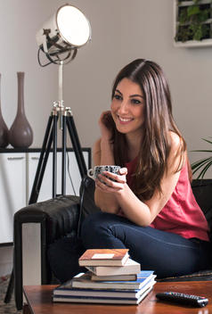 A beautiful brunette sits in her apartment drinking coffee and smiling while touching her hair