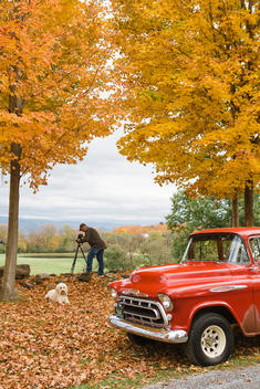 Older white man making photographs on a tripod of peak fall foliage color with a large white dog and classic red pick-up truck on dirt road.