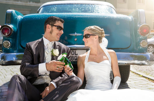 Bridal couple sitting in front of vintage car toasting with piccolo