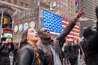 A black African-American points to a Jumbo-Tron with his girlfriend beside him amidst a crowd of tourists and the US Armed Forces Recruiting Station behind in Times Square. New York, NY
