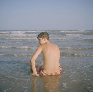 A teenage boy looking over his shoulder relaxes in the water at the edge of the ocean at the beach in Galveston, Texas