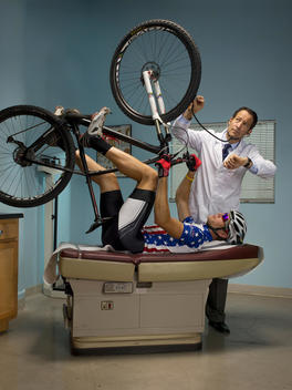 A bicyclist lies on his back on a table in an exam room holding his bike in the air while a doctor uses a stethoscope on the front tire
