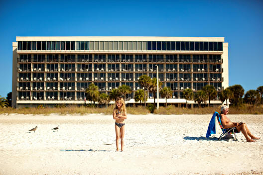 A Young Girl Stands On The Beach In Front Of A Large Hotel, With An Old Man Reading The Newspaper Next To Her.