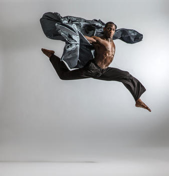 Black male dancer jumping like a gazelle with grey metallic prop looking forward against white