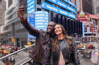 An interracial couple smile while taking a selfie with an Android phone with a crowd of tourists on street below in Times Square. New York, NY