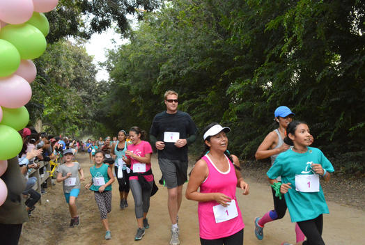 Runners of varied ages and ethnic groups approach the finish line in a 5K race on a tree-lined dirt road, with a tall man towering over the women.
