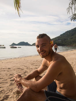 An athletic man with a Mohawk sitting on the beach smiling with a flower behind his ear.
