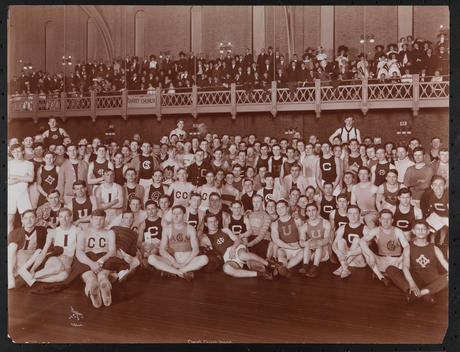 Group Of Young Men In Athletic Gear Standing On The Gymnasium Floor Of Christ Church With Band And Spectators.