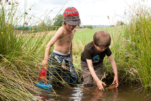 Two boys are playing with little wooden boats by a stream. They are standing in the water knee deep. The background consists of fields and trees.