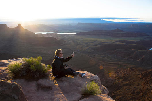 At sunrise a man sits on the edge of a rock to get a better view of the American West landscape as he takes pictures with his I Phone.