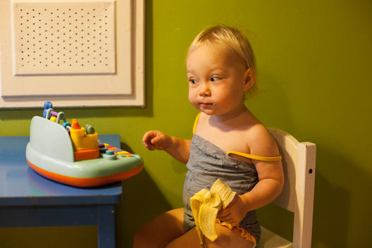 A blonde toddler girl in bathing suit take a big bite of a banana as she sits in a small chair at a small blue table with the other in front of a small toy.