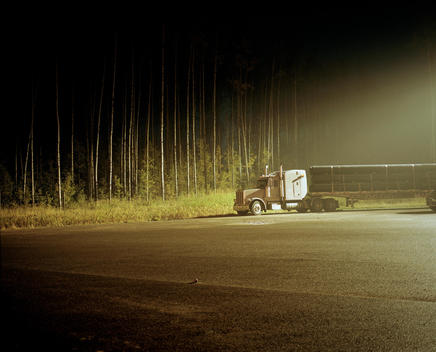 A semi truck carrying pipes is parked under the light of a street lamp in an empty parking lot.