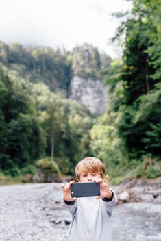 6 year old blonde boy in gray shirt taking selfie in alpine landscape with creek behind him.