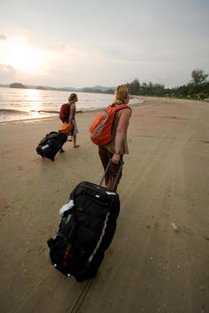 Two females make their way across the sand with rollerbags in town. Searching for the perfect hut on the beach in Thailand.