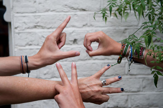 Two pairs of hands are spelling out \'Love\'. The arms are graced with bracelets. A white wall in the background.