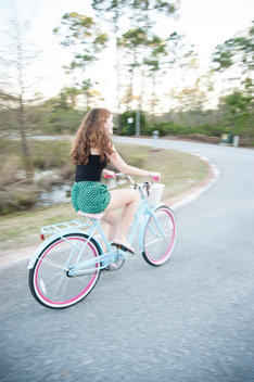 A teenage caucasian girl, long brown curly hair, riding a blue and pink cruiser bicycle down street.