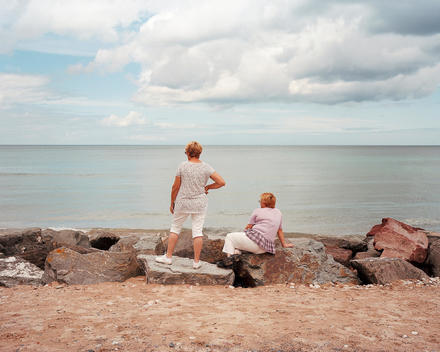 Two Women Looking Out To Sea, One Sitting On Rocks And One Standing