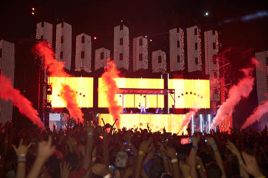 A crowd at a concert with a light show and dj booth in the background.