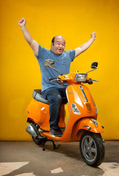 A chubby, bald man is sitting on an orange electric scooter with both arms up in the air in front of a yellow background