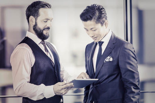 caucasian businessman with beard and asian businessman standing in office and talking while holding a tablet computer