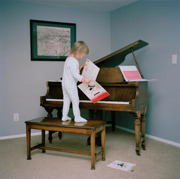 A young blonde girl wearing blue heart pajamas plays on the piano standing on the bench and throwing music sheets around