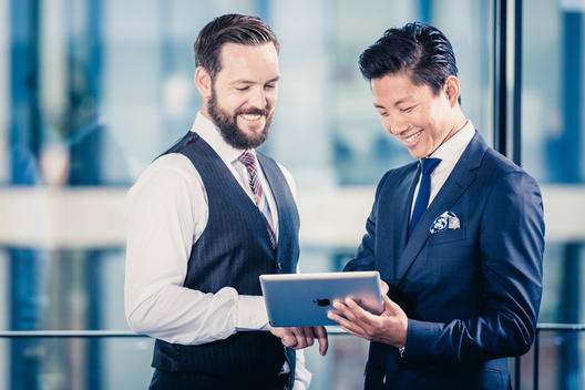 caucasian businessman with beard and Asian businessman standing in office and talking while holding a tablet computer