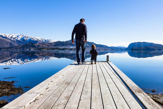 Father and son walking by a calm Fjord with snow covered mountains in the distance, Myking, Norhordaland, Norway.