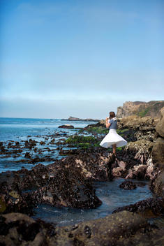 Woman in white skirt stands on rocks looking out to the sea.