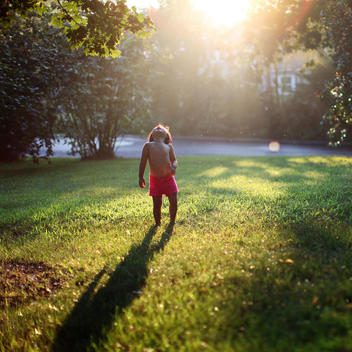 A shirtless young child, girl, wearing pink shorts stands under a tree after a summer afternoon rain storm looking up at the sky while tiny raindrops fall on her face.