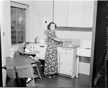View Of A Woman Cooking At The Stove; Baby In A Highchair By A Table.