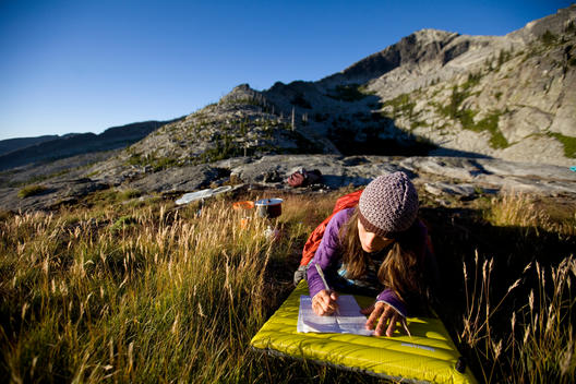 Adult woman laying, writing, on a camp pad in the mountains.