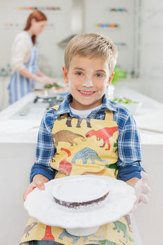 Boy covered in flour holding cake in kitchen