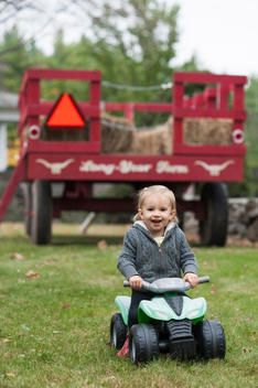 Toddler rides toy ATV behind hay wagon on a farm.