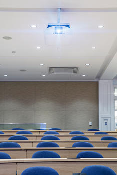 Auditorium style, lecture class room with rows of chairs and long wood tables with projector hanging from ceiling.